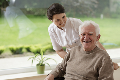 CNA standing behind elderly man in wheelchair next to a window